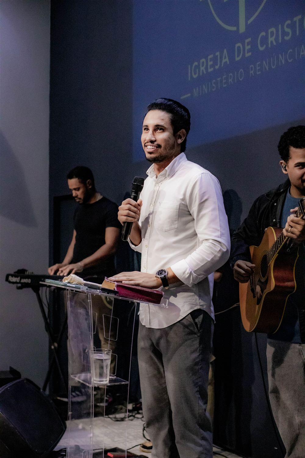 Pastor Kaic sorrindo no púlpito, com a Bíblia aberta e o microfone na mão